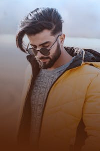 Young man with a beard and sunglasses wearing a yellow jacket outdoors.